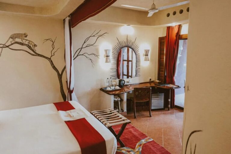 A room with a luggage rack, a desk, a kettle and a small fridge, under a ceiling fan