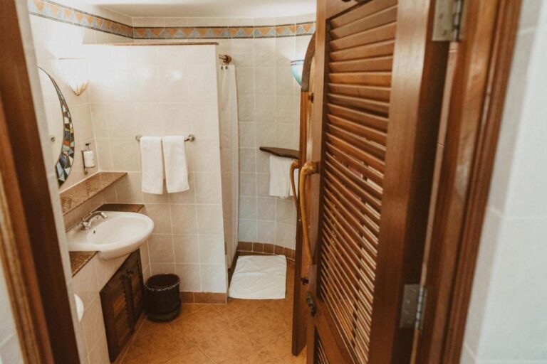A bathroom seen from its louvered wooden door, with a sink and a shower with a curtain