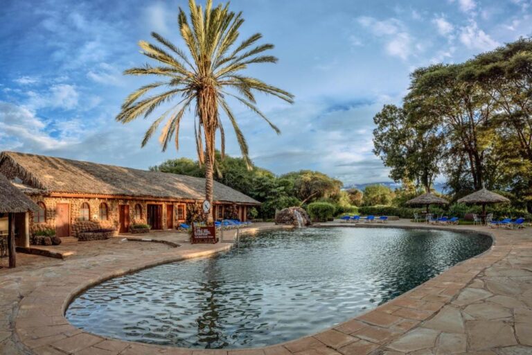 The pool at Amboseli Sopa Lodge, with a palm tree, thatched umbrellas and a stone building
