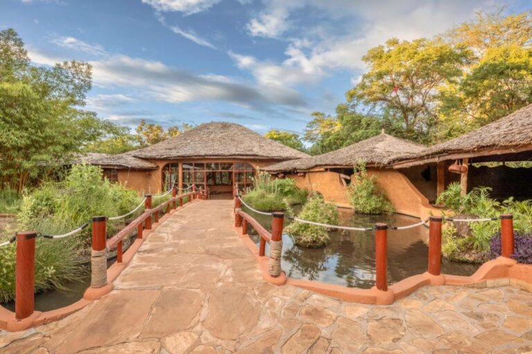 A rope-lined stone walkway crossing a pond, toward thatched roofs