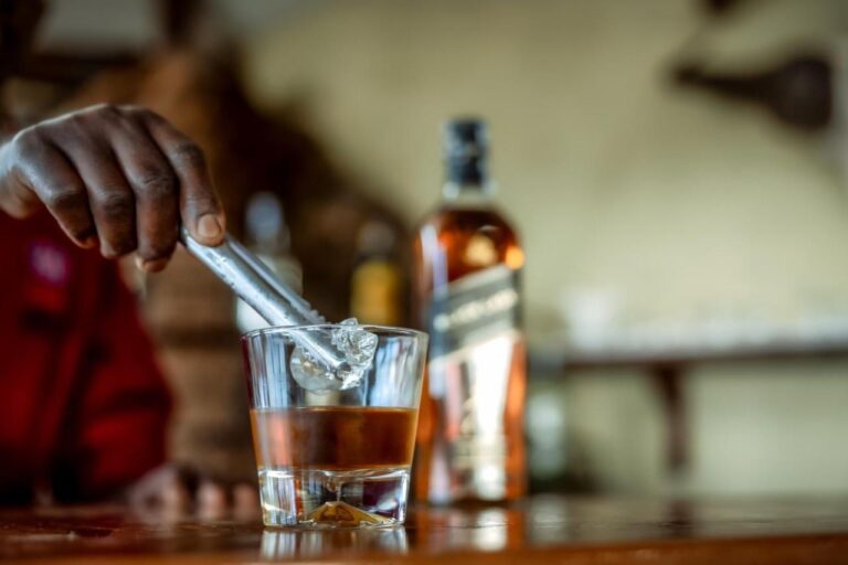 A hand dropping an ice cube into a glass of whisky, on the bar counter