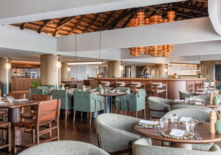 A restaurant with green armchairs in front of a long bar, under orange pendant lights