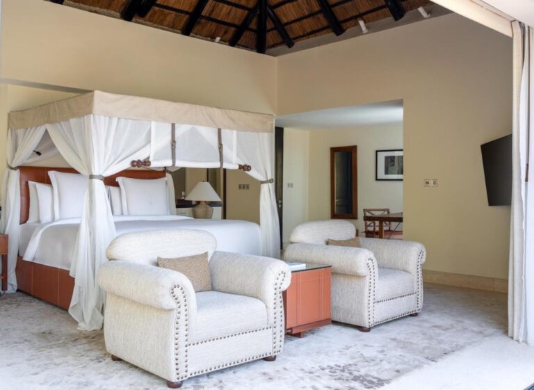 A room with a four-poster bed and two white tufted armchairs