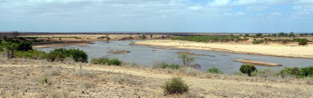 The Galana River, wide and brown, curves between pale sandbanks and bushes, under a hazy sky