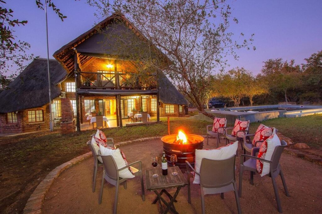 The brick and thatch building of Imbube Safari Lodge at dusk, with a lit fire pit surrounded by armchairs, near the pool