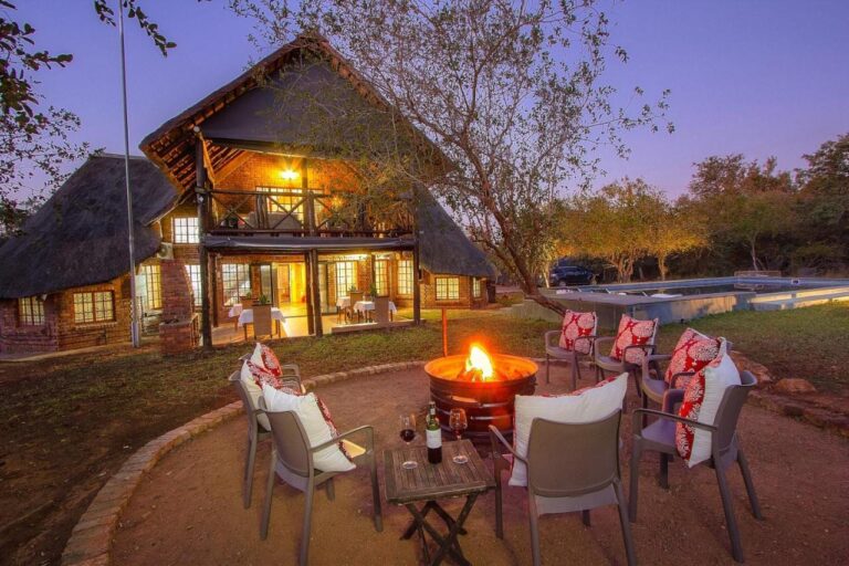 The brick and thatch building of Imbube Safari Lodge at dusk, with a lit fire pit surrounded by armchairs, near the pool