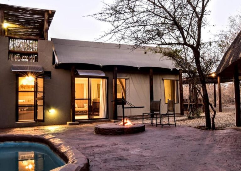A tent at Jackalberry Ridge lit up at dusk, with its private pool, a fire burning in the fire pit and two chairs on the deck