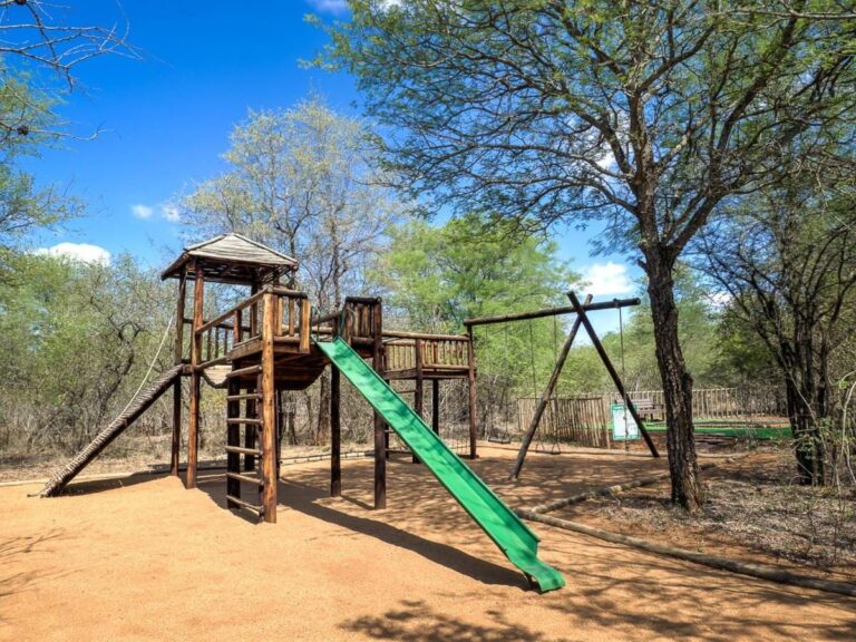 The children's playground, with a wooden tower, a green slide and a swing, under the trees