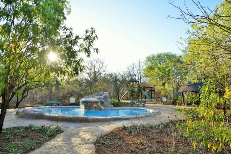 A pool with a rock waterfall, in front of the playground and a thatched shelter, backlit
