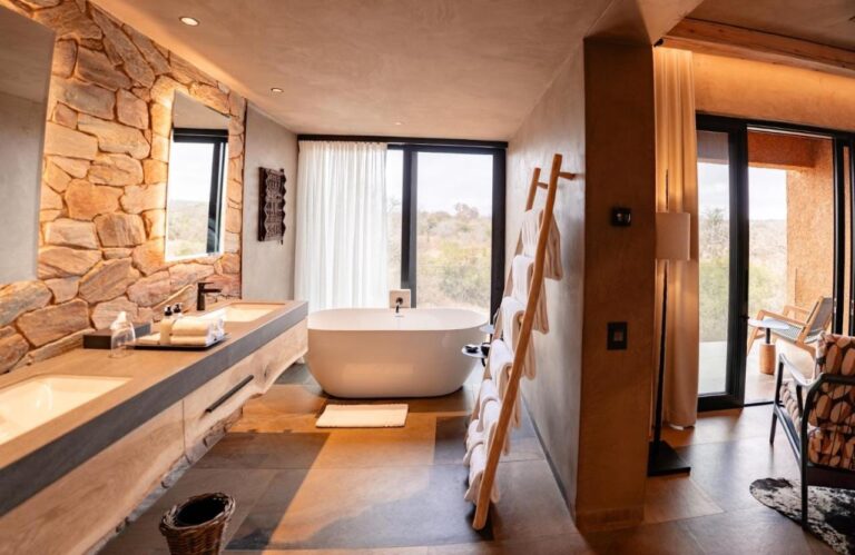The bathroom of a suite, with a bathtub in front of the window and a double sink against a stone wall