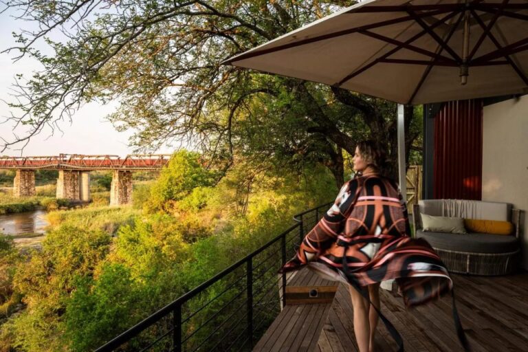 A woman in a bathrobe on a wooden deck, under a parasol, facing the bridge and the river