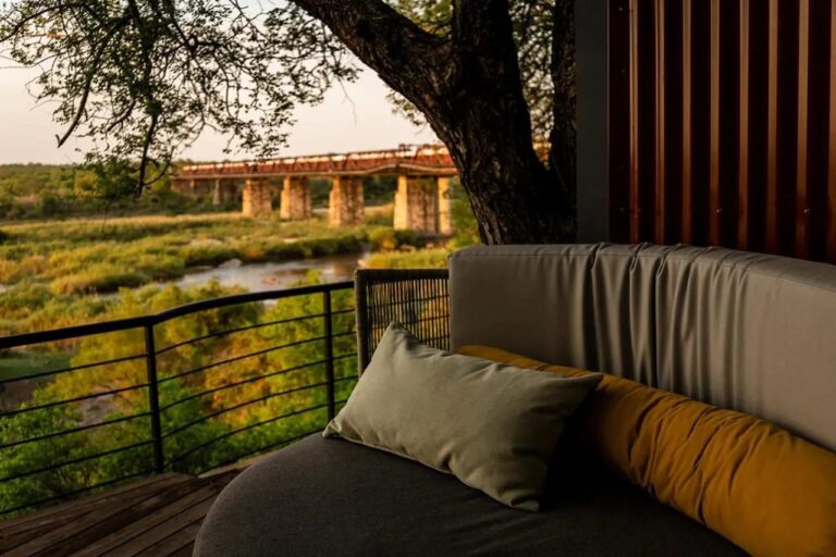 A curved bench on a terrace, under a large tree, facing the bridge and the river