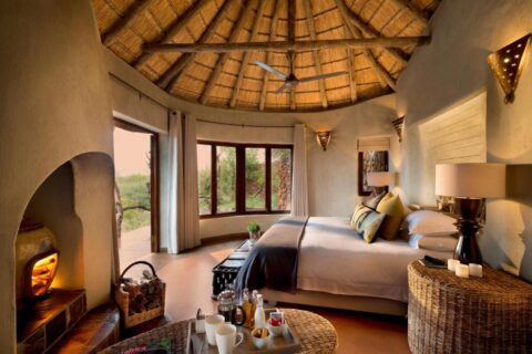 A suite at Madikwe Safari Lodge under a thatched roof, with a double bed and a lit fireplace