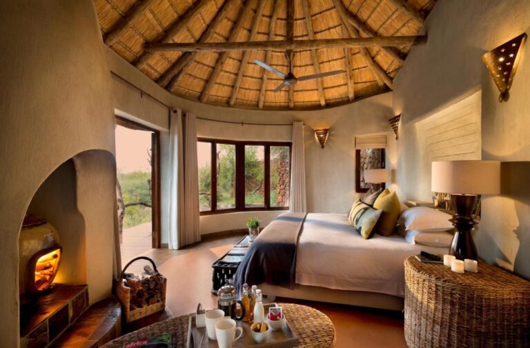 A suite at Madikwe Safari Lodge under a thatched roof, with a double bed and a lit fireplace