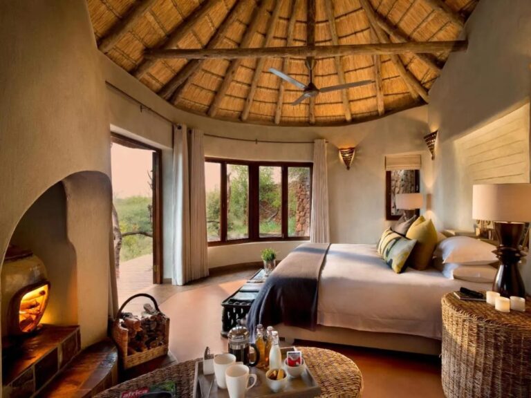 The double bed of a suite under the thatched roof frame, between the fireplace and the windows over the bush