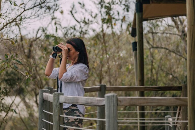 A woman with binoculars on the wooden deck of a tent, facing the bush