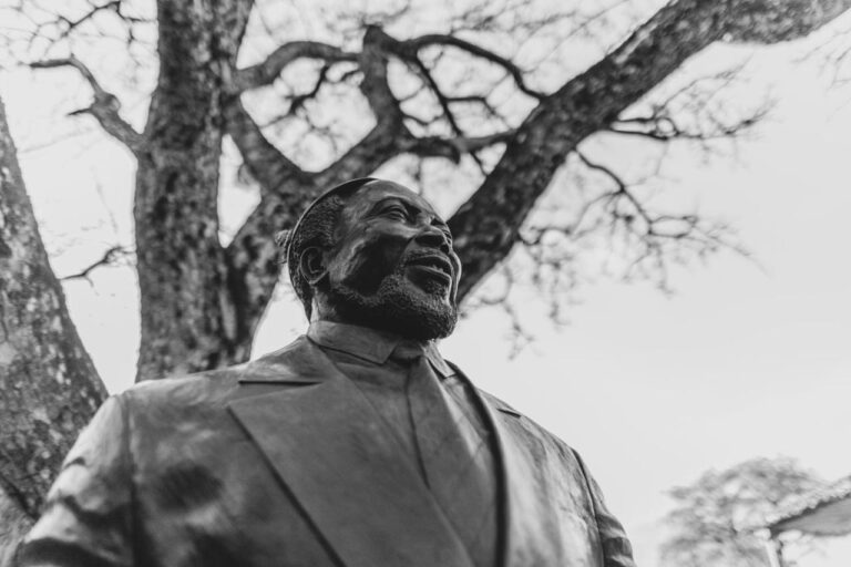 The bronze statue of a smiling bearded man in a suit, under a tree, in black and white
