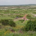 View from Observation Hill over a green plain patched with salt, a red track, three 4x4s parked near a small ocher building, low hills in the distance