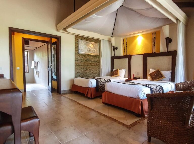 A twin room under a canvas canopy, with a rattan armchair and the bathroom door open