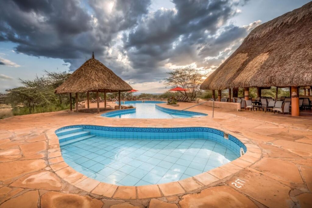 A small tiled pool in the foreground and the large pool of Samburu Sopa Lodge, between a thatched shelter and a terrace with a thatched roof