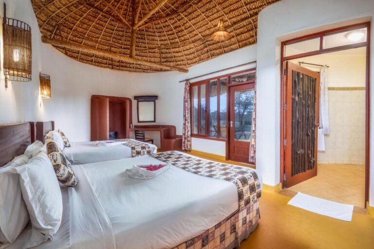 A twin room under a thatched ceiling, with a carved wooden door open onto the bathroom