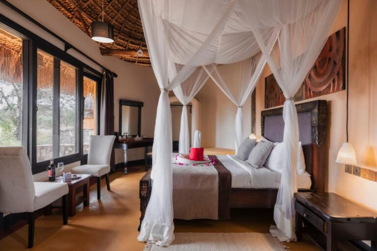 A room with a four-poster bed draped in white mosquito nets, and two armchairs in front of the large windows