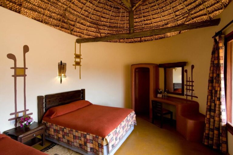 A room with a large bed under an orange cover, beneath a thatched ceiling, with a curved ocher dressing table