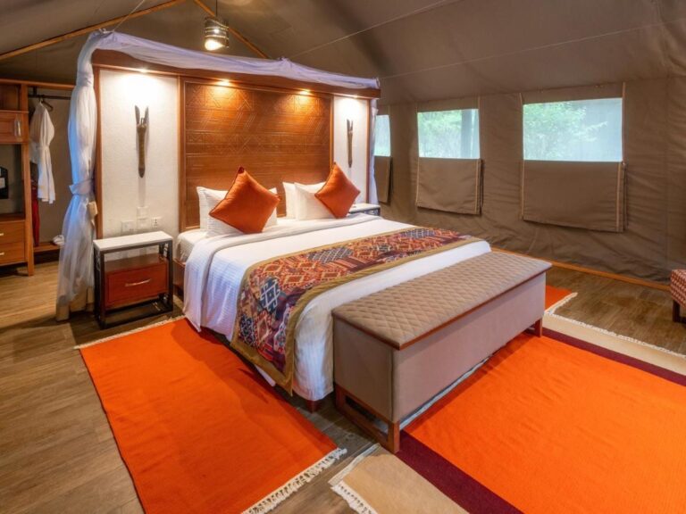A tent with a large bed under a mosquito net, orange cushions and a bench at the foot of the bed