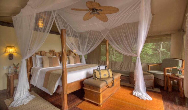 A carved wooden four-poster bed under a mosquito net, in a tent with windows facing the trees