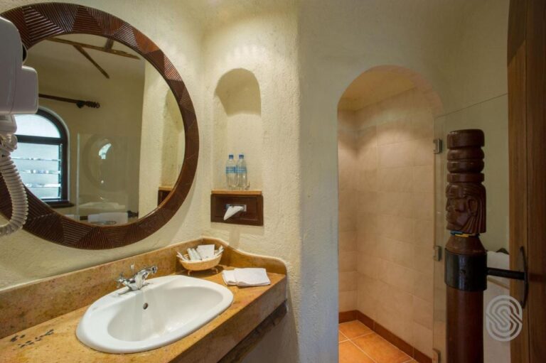 A bathroom with stucco walls, a round mirror framed in carved wood and a shower under an arch