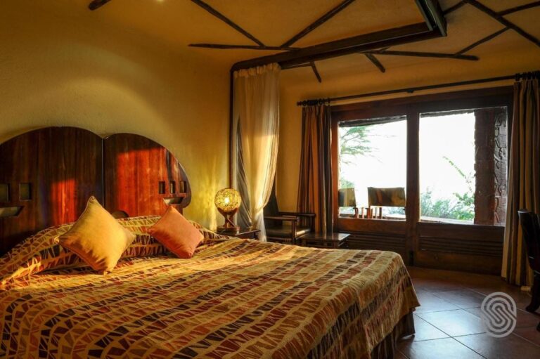 A double bed with an orange and brown patterned bedspread, and two armchairs on the balcony behind the glass