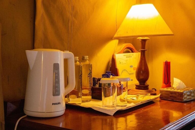 A kettle, bottles of water and glasses on a desk, under a lamp