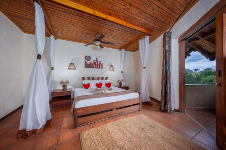 A room with a double bed under a wooden ceiling, with sheer curtains and a balcony