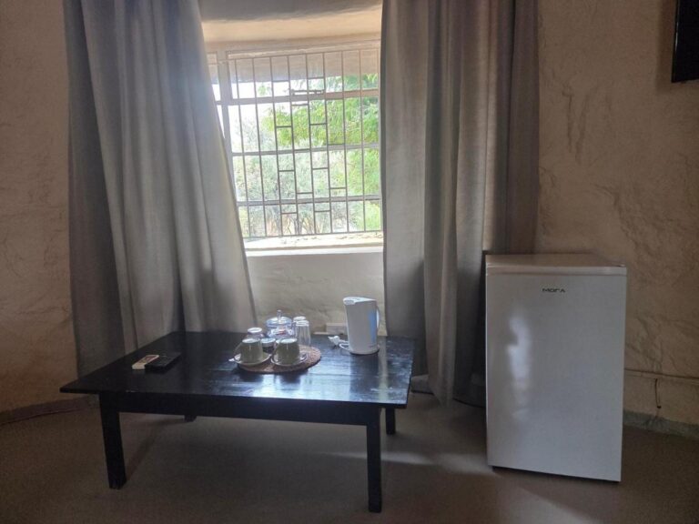 A coffee table with cups and a kettle in front of a screened window, next to a small fridge