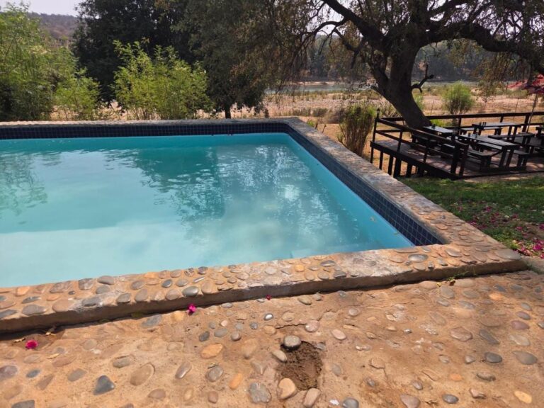 The pebble-edged pool of Three Bridges B&B, in front of a wooden deck with picnic tables, and the river beyond