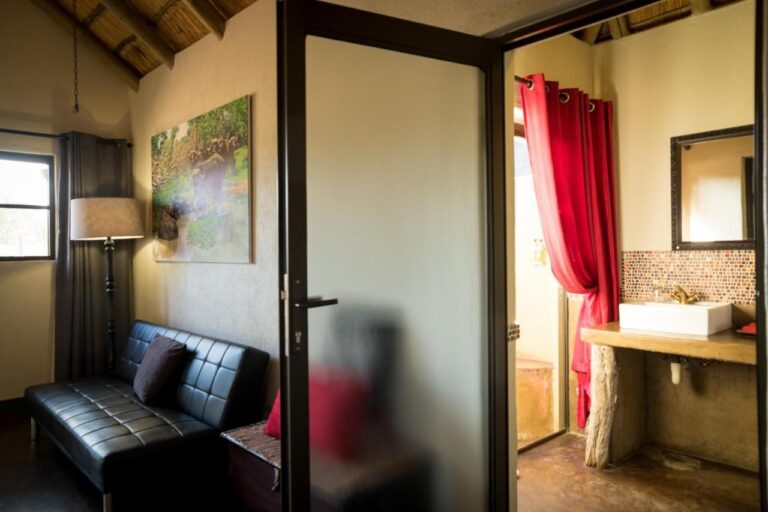 The seating area of a room, with a black leather sofa, and the bathroom behind a glass door